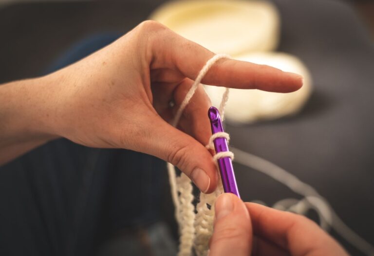 Hand hält eine lila Häkelnadel und legt den Faden um den Finger beim Häkeln